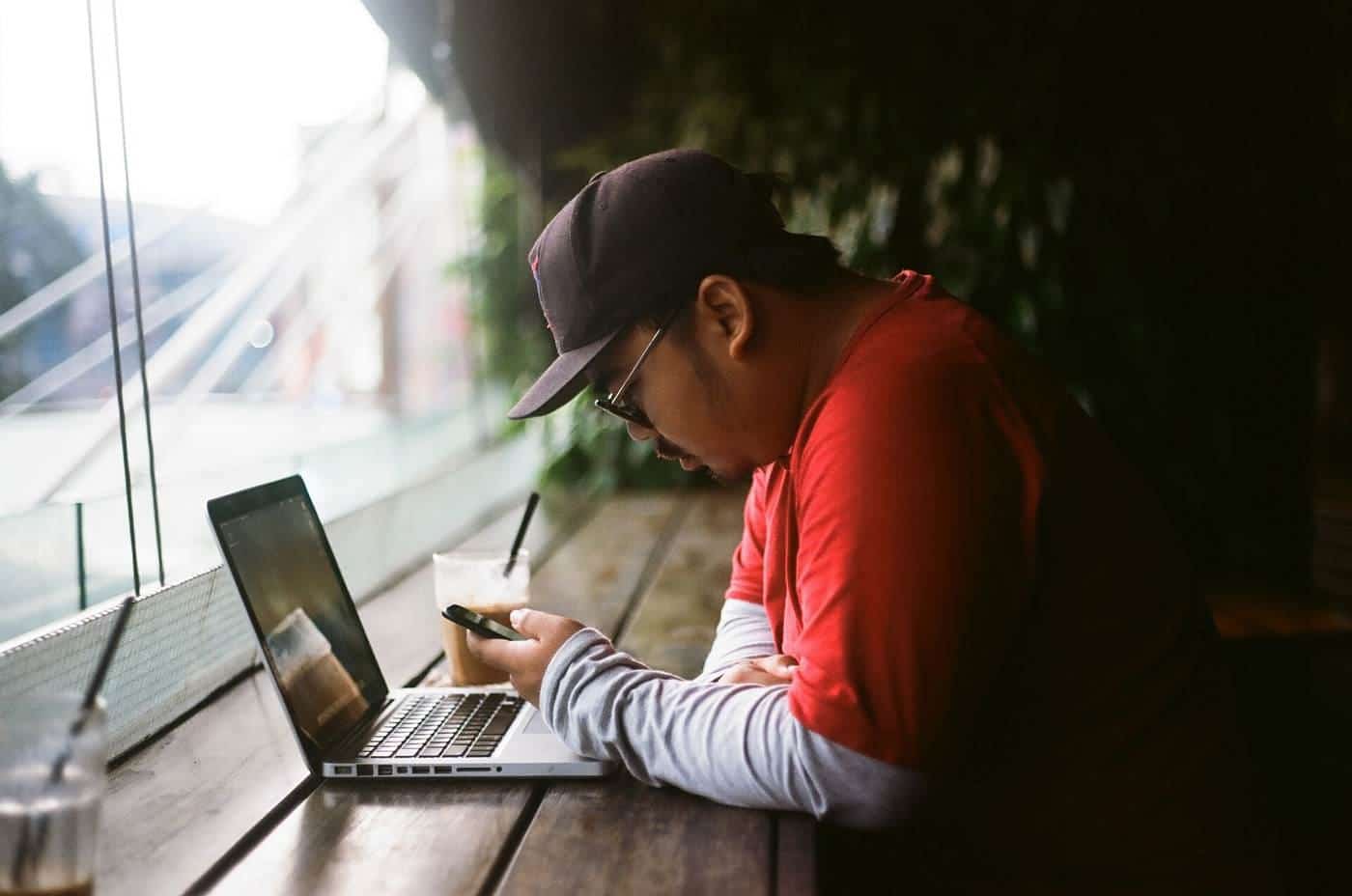 A man in a cap working on a laptop at a table by a window